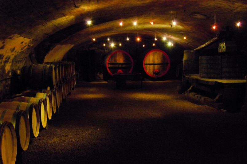 Cave à vin authentique : fûts de chêne et pressoir ancien Cave à vin voûtée avec tonneaux en chêne, pressoir ancien et fûts éclairés dans une ambiance tamisée et authentique.